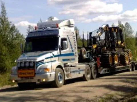 Lastbil som transporterar en skogsmaskin på trailer längs en skogsbilväg.